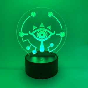 May include: A green LED night light with a white outline of the Sheikah Eye from The Legend of Zelda: Breath of the Wild. The light is on a black base with a power switch.