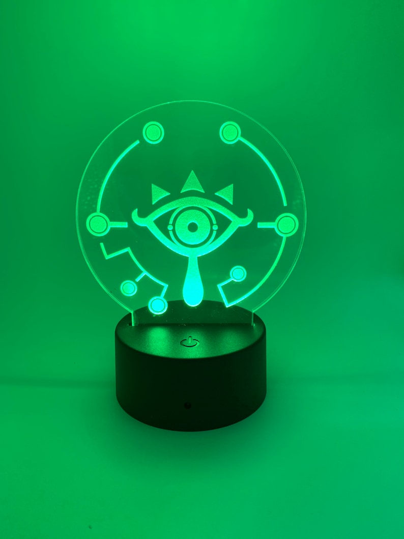 May include: A green LED night light with a white outline of the Sheikah Eye from The Legend of Zelda: Breath of the Wild. The light is on a black base with a power switch.