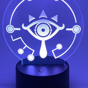 May include: A white LED night light shaped like a circle with a stylized eye design inside. The eye has a white outline and a black pupil. The light is on a black base with an on/off switch.