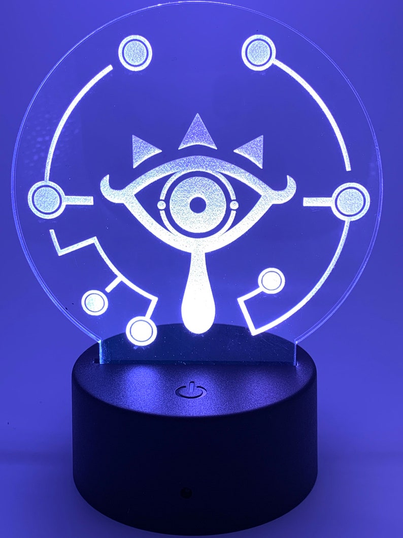May include: A white LED night light shaped like a circle with a stylized eye design inside. The eye has a white outline and a black pupil. The light is on a black base with an on/off switch.