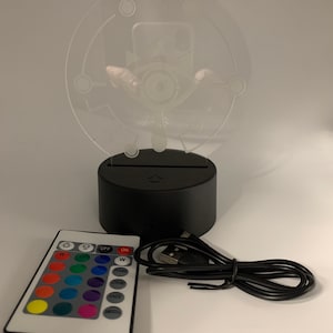 May include: A black and clear night light with a white design of an eye with a crown on top. The light is on a black base with a USB port and a remote control with 16 color options, "OFF", "ON", "W", "FLASH", "STROBE", "FADE", and "SMOOTH" buttons.