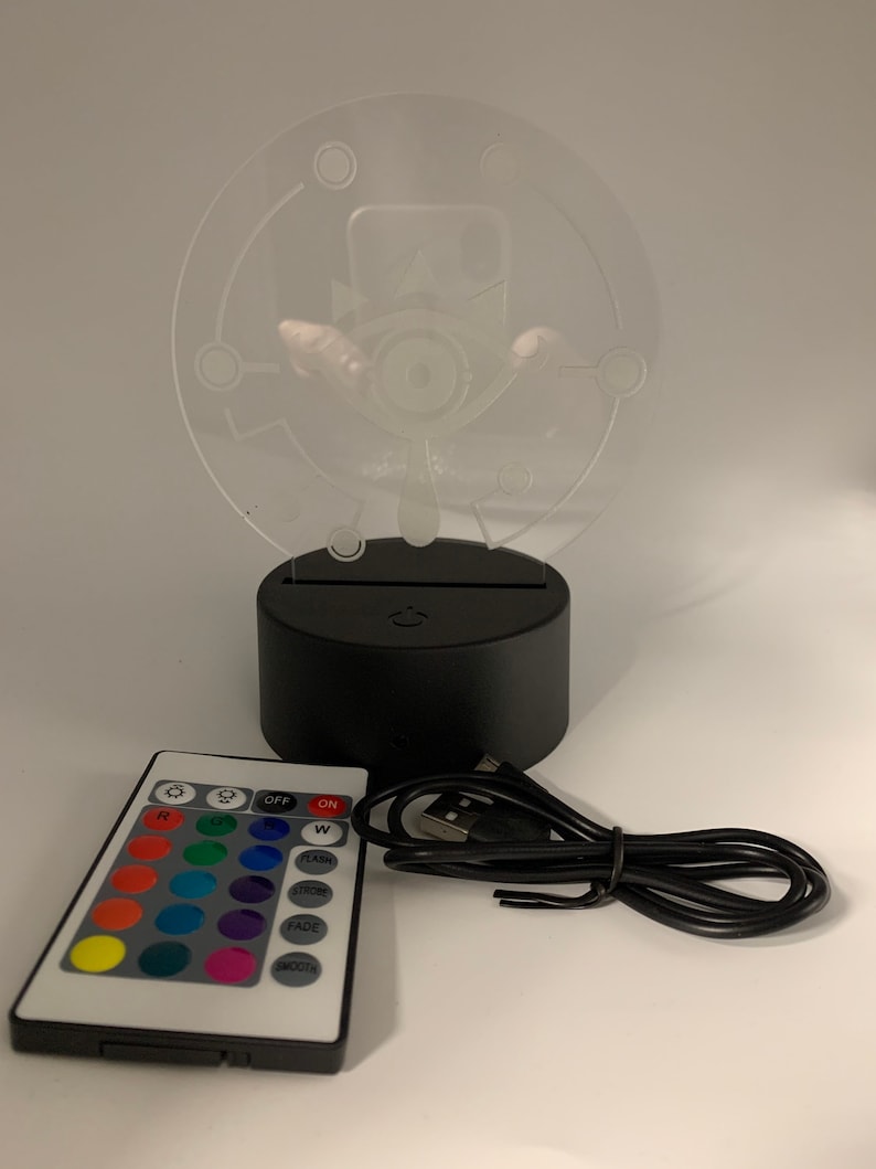 May include: A black and clear night light with a white design of an eye with a crown on top. The light is on a black base with a USB port and a remote control with 16 color options, "OFF", "ON", "W", "FLASH", "STROBE", "FADE", and "SMOOTH" buttons.