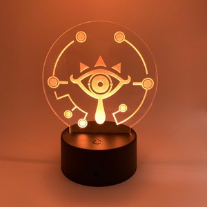 May include: A clear acrylic night light with a white outline of the Sheikah Eye symbol from the Legend of Zelda video game series. The light is on a black base with a switch.