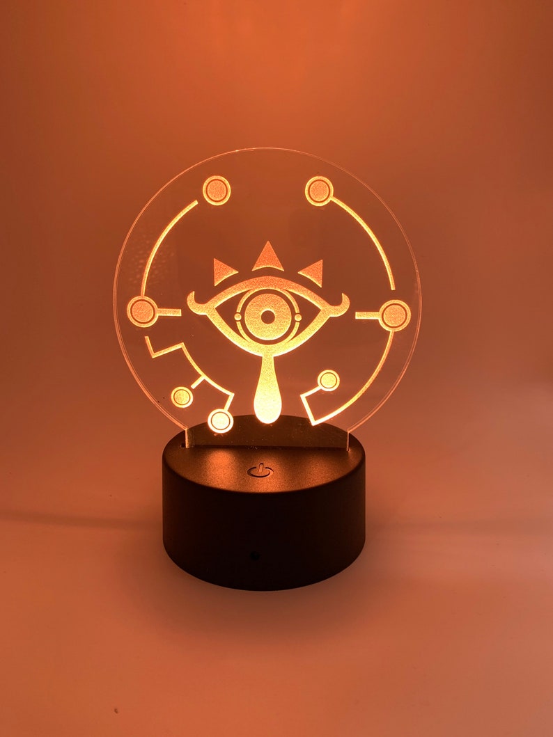May include: A clear acrylic night light with a white outline of the Sheikah Eye symbol from the Legend of Zelda video game series. The light is on a black base with a switch.