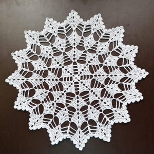 Könnte beinhalten: Weißes, handgefertigtes Häkeldeckchen mit Sternmuster. Das komplizierte Spitzendesign weist ein sich wiederholendes Motiv aus stilisierten Blättern und offenen Räumen auf, wodurch ein zartes und luftiges Aussehen entsteht. Das Deckchen ist kreisförmig mit einem gewellten Rand.