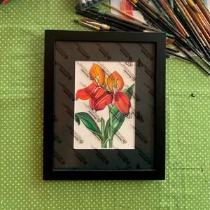 Pode incluir: Uma pintura de aquarela emoldurada de uma flor vermelha e laranja com folhas verdes. A pintura está em uma moldura preta com um passe-partout branco.
