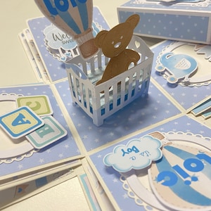 May include: A blue and white explosion box with a teddy bear in a crib, a hot air balloon, and the words "It's a Boy!" and "Welcome Sweet One".