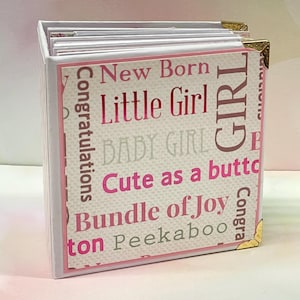 Puede incluir: Álbum de fotos blanco con texto rosa y gris en la portada. El texto dice "Felicidades", "Recién nacido", "Niña pequeña", "Bebé niña", "Lindo como un botón", "Paquete de alegría", "Asomarse" y "Chica".