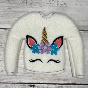 May include: White elf-sized sweater with a unicorn design embroidered on the front. The unicorn has a gold horn, pink ears, and purple flowers around its head.