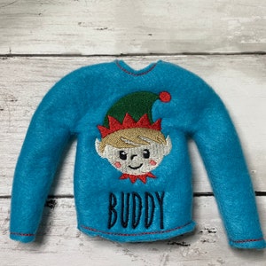 May include: Blue elf shirt with a green hat and red trim. The shirt has the word "BUDDY" embroidered on the front.