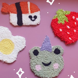Puede incluir: Una colección de cuatro artículos decorativos hechos a mano, coloridos y texturizados. Incluye una pieza de sushi, una fresa roja, un huevo frito y una rana verde con un cuerno morado. Cada artículo tiene un diseño único y una cara sonriente.