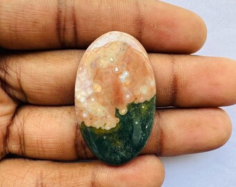 May include: An oval-shaped gemstone with a mix of colors. The top portion is a light beige with white spots, transitioning to a peachy-pink hue. The bottom half is a deep green. The stone is held in a hand.