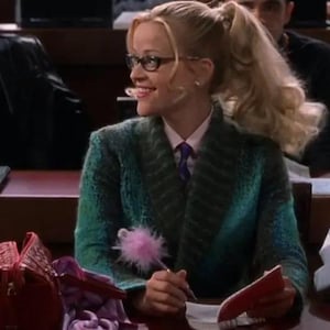 Op de afbeelding: Een persoon met blond haar in een paardenstaart draagt een getextureerde blauwgroene blazer met sjaalkraag over een lichtroze overhemd met kraag en een paarse das. Ze draagt een bril met zwart montuur en houdt een pen met roze pompon vast.