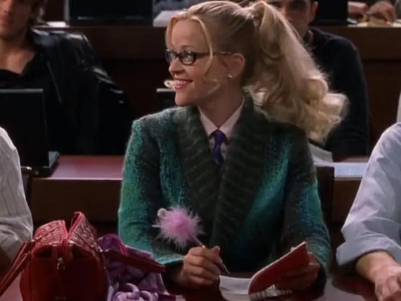 Op de afbeelding: Een persoon met blond haar in een paardenstaart draagt een getextureerde blauwgroene blazer met sjaalkraag over een lichtroze overhemd met kraag en een paarse das. Ze draagt een bril met zwart montuur en houdt een pen met roze pompon vast.