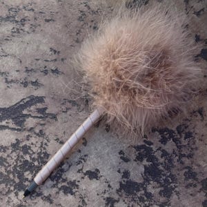 Op de afbeelding: Een decoratieve pen met een pluizige, lichtbruine verenbol. De pen heeft een schacht die is omwikkeld met een bijpassend lichtbruin materiaal. De pen heeft een zwarte punt en ligt op een getextureerde achtergrond.