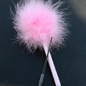 Op de afbeelding: Een balpen met een grote, pluizige lichtroze veren topper. De penhouder is omwikkeld met een glad, bijpassend lichtroze satijnen lint en de schrijfpunt is zichtbaar met een donkergrijze plastic behuizing aan de onderkant.