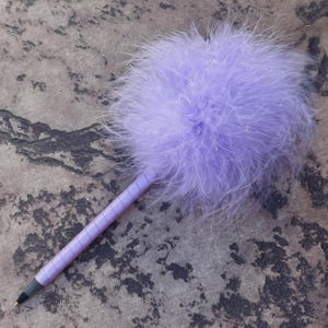 May include: A lavender pen with a fluffy feather ball on top. The pen's barrel is a matching lavender color with a ribbed design. The pen is set against a mottled gray and brown surface.