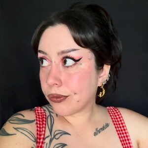 May include: A person with dark hair and makeup, including pink eyeshadow and black eyeliner, is wearing gold crescent moon earrings. The person is wearing a red and white checkered top. A tattoo is visible on the person's shoulder.