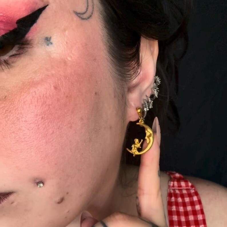 May include: Gold-colored moon and stars earrings with a dark rectangular backing. The earrings feature a crescent moon design with a figure sitting on it. A silver-colored ear cuff with star-shaped accents is also visible.