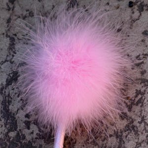 Op de afbeelding: Een lichtroze veren pompon bevestigd aan een slank, bijpassend lichtroze handvat. De bolvormige pompon bestaat uit talloze zachte, ijle veren die in alle richtingen uitsteken, wat zorgt voor een volumineus en getextureerd uiterlijk.