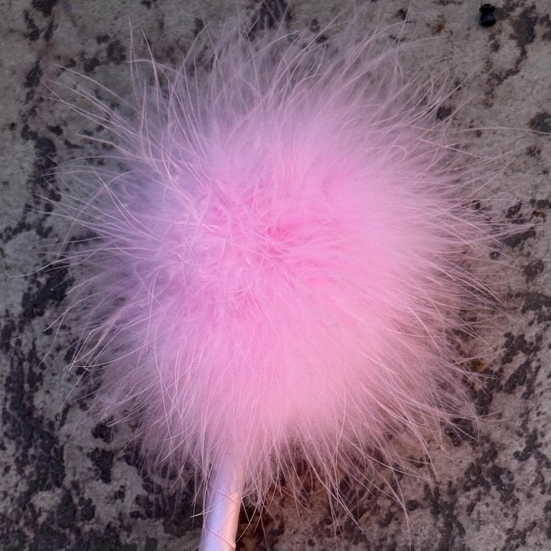 Op de afbeelding: Een lichtroze veren pompon bevestigd aan een slank, bijpassend lichtroze handvat. De bolvormige pompon bestaat uit talloze zachte, ijle veren die in alle richtingen uitsteken, wat zorgt voor een volumineus en getextureerd uiterlijk.