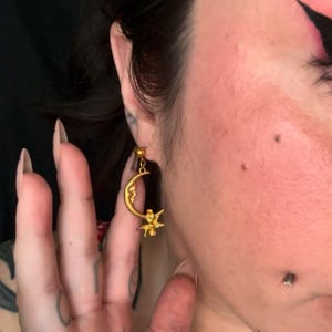 May include: Gold-colored dangle earrings. Each earring features a crescent moon with a smiling face and a small figure of an angel. The earrings are attached to a small gold ball that connects to the ear.