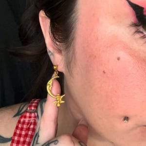 May include: Gold-colored celestial-themed earring featuring a crescent moon with a face and a star. The earring has a small ball at the top for the ear piercing. The earring is dangling from the ear.