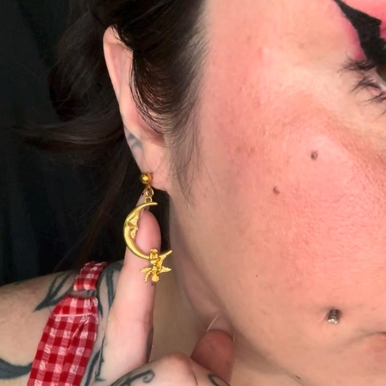 May include: Gold-colored celestial-themed earring featuring a crescent moon with a face and a star. The earring has a small ball at the top for the ear piercing. The earring is dangling from the ear.