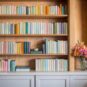 May include: A wooden bookshelf with multiple shelves filled with colorful books. The bookshelf is in a room with a window and a vase of pink flowers on a white countertop.