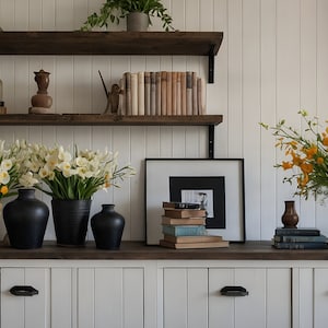 May include: A white wall with two wooden shelves holding books, vases, and decorative items.  The shelves are above a white cabinet with three drawers.  There are three vases with white flowers and one vase with yellow flowers.