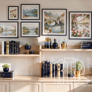 May include: A gallery wall featuring framed watercolor landscape paintings in black frames. The paintings depict scenes of European towns, bridges, and waterways. The display includes books, plants in gold and navy pots, and decorative vases on wooden shelves.