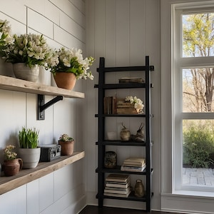 May include: A white wall with two wooden shelves with black metal brackets. The shelves are decorated with potted plants, a small black purse, and a few other items. A black ladder shelf with five shelves is on the right side of the image. The ladder shelf is filled with books and decorative items. A large window with white trim is on the right side of the image. The window looks out onto a green lawn and trees.