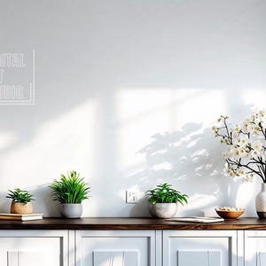 Puede incluir: Una pared blanca con un logotipo blanco que dice "RTP Digital Art Studio". Hay una lámpara blanca con una pantalla blanca sobre una mesa con un jarrón blanco con flores blancas, una tabla de cortar de madera y tres plantas en maceta. La mesa está delante de un armario blanco con tres cajones.