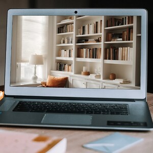 May include: A laptop computer displaying a white bookshelf filled with books. The bookshelf is in a room with a window and a lamp.