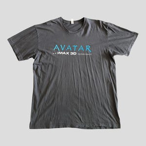 May include: Dark gray t-shirt with the word "AVATAR" in blue, above "AN IMAX 3D EXPERIENCE" in white. The shirt is a crew neck style and appears to be made of a soft material. The shirt is laid flat on a light gray surface.