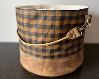 May include: A cylindrical storage basket featuring a black and brown buffalo check pattern on the upper portion and a solid tan base. The basket includes a tan handle attached with brass-colored metal hardware and a light-colored interior lining.