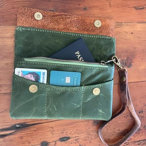 Puede incluir: Cartera de lona verde con solapa y correa de muñeca de cuero marrón. La cartera está abierta, revelando un pasaporte, tarjetas de crédito y un bolsillo con cremallera. La cartera se asegura con cierres de presión de latón.