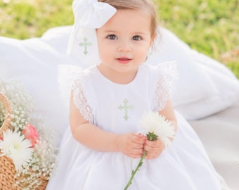May include: A white dress featuring lace flutter sleeves and a light green embroidered cross on the bodice. The outfit includes a matching white hair bow with a light green embroidered cross, and a white flower held in the hands.