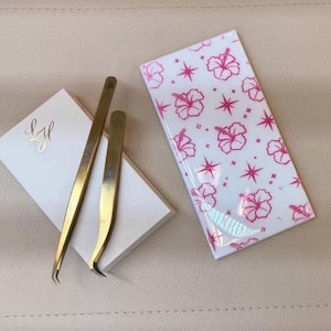 May include: Gold-colored eyelash extension tweezers and a white box with gold lettering, next to a white case with a pink floral pattern. The tweezers are curved and straight. The case has a glossy finish.