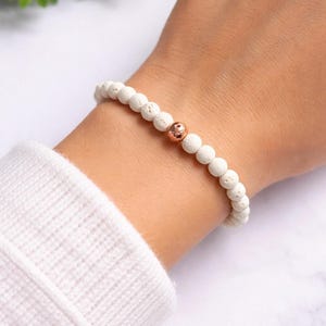 May include: White beaded bracelet with a single copper-colored bead. The bracelet is made of small, round, textured white beads. The copper bead is centered on the bracelet. The bracelet is worn on a wrist, with a white sweater sleeve visible.