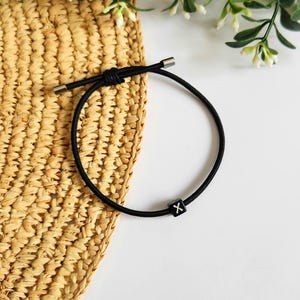 May include: A black cord bracelet with a small, square bead displaying the letter "X". The bracelet is set against a white backdrop, with a woven straw texture and foliage in the background.
