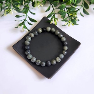 May include: A bracelet with grey and black beads sits on a square black dish. The bracelet has a mix of round, grey-green beads and darker, textured beads. The background includes green foliage and white flowers.