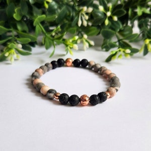 May include: A beaded bracelet featuring a mix of natural stone beads in shades of gray, pink, and black. Accented with copper-colored beads. The bracelet is displayed on a white surface with greenery in the background.