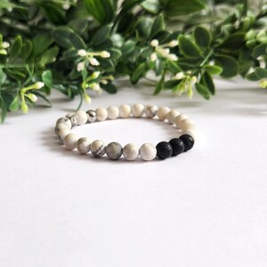 May include: A beaded bracelet featuring a mix of white, gray, and black round beads. The bracelet is made of natural stone beads and is displayed on a white surface with green foliage in the background. The bracelet is a stylish accessory.