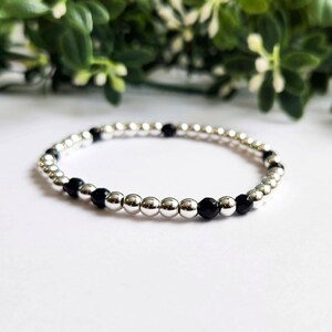 May include: A silver beaded bracelet featuring alternating silver and black beads. The bracelet is composed of small, round beads strung together. The background is white with green foliage.