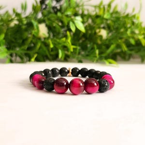 May include: A bracelet with alternating black and magenta beads. The bracelet includes lava rock beads and polished, round magenta beads. The bracelet is displayed on a white surface with green foliage in the background.