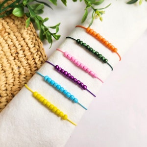 May include: Six colourful beaded bracelets on a white surface. The bracelets are made with small, round beads in shades of yellow, blue, purple, pink, green, and orange.