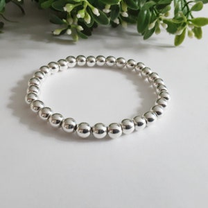 May include: A silver beaded bracelet. The bracelet features numerous small, round, shiny silver beads strung together. The beads are uniform in size and shape, forming a smooth, continuous loop. The background is white with green foliage.