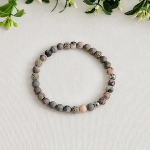 May include: A bracelet made of small, round beads in shades of grey, brown, and pink. The bracelet includes textured beads and is displayed on a white surface. Green foliage with small white flowers is visible in the background.