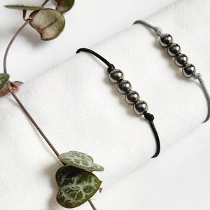 May include: Two bracelets with black and grey cords and silver beads. The bracelets are on a white surface.